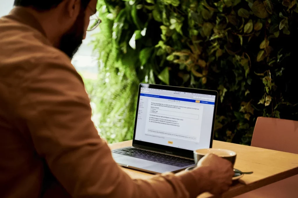 Man sits at a wooden table using a laptop, with a green plant wall in the background. He holds a cup, creating a calm, focused atmosphere.