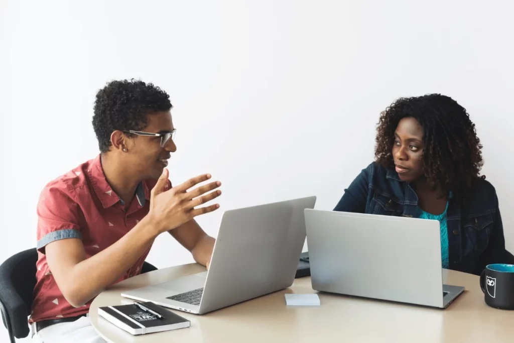 Two people sit at a table with laptops, engaged in a discussion. One gestures expressively while the other listens attentively. The setting is a bright office.