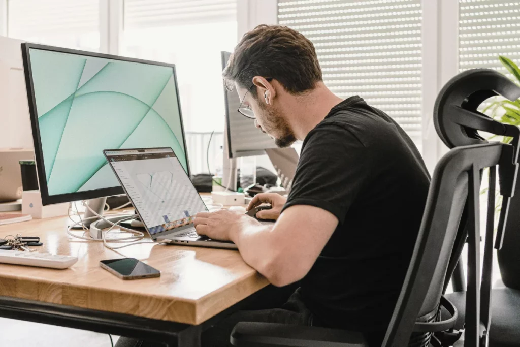 Man focused on laptop with smartphone and monitor on desk.