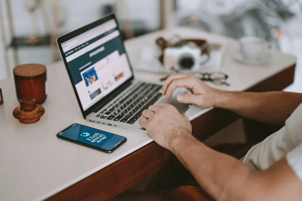 Man using laptop while smartphone rests on white desk.